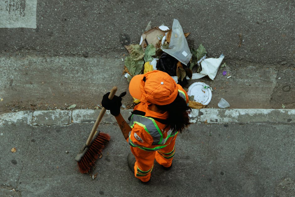 A street cleaning scene showing a worker in a high-visibility green and yellow uniform with a white hard hat, operating a street sweeper machine on a paved road near a red brick wall and green hedge.